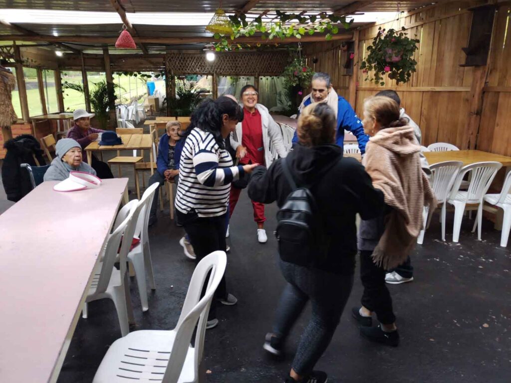 Grupo de residentes adultos mayores bailando en ronda y disfrutando junto al equipo de cuidadoras durante un paseo a una hacienda.