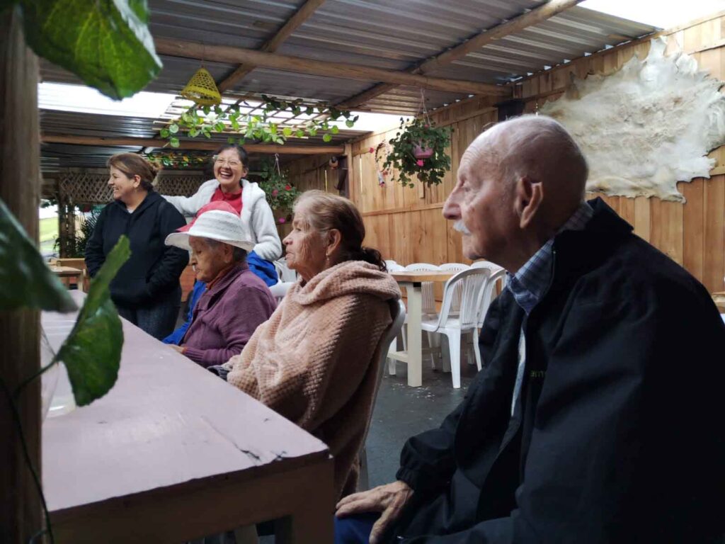 Grupo de residentes adultos mayores sentados junto a personal de enfermería conversando durante paseo a una hacienda mientras esperan el almuerzo.