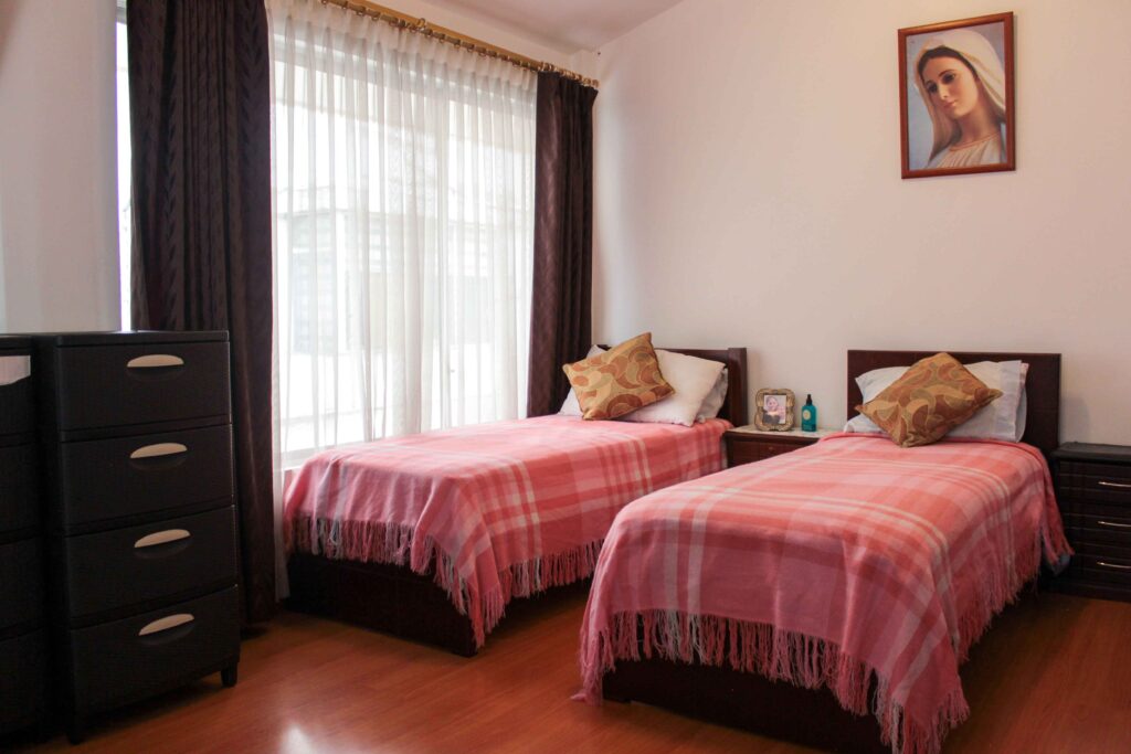 Habitación compartida de residencia geriátrica con camas, mesas auxiliares y mobiliario.