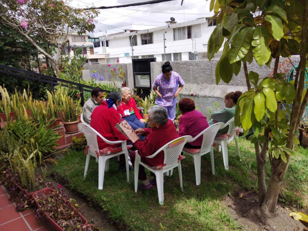 Adultos mayores residentes leyendo al aire libre y conversando acompañados por personal de la residencia en el jardín.