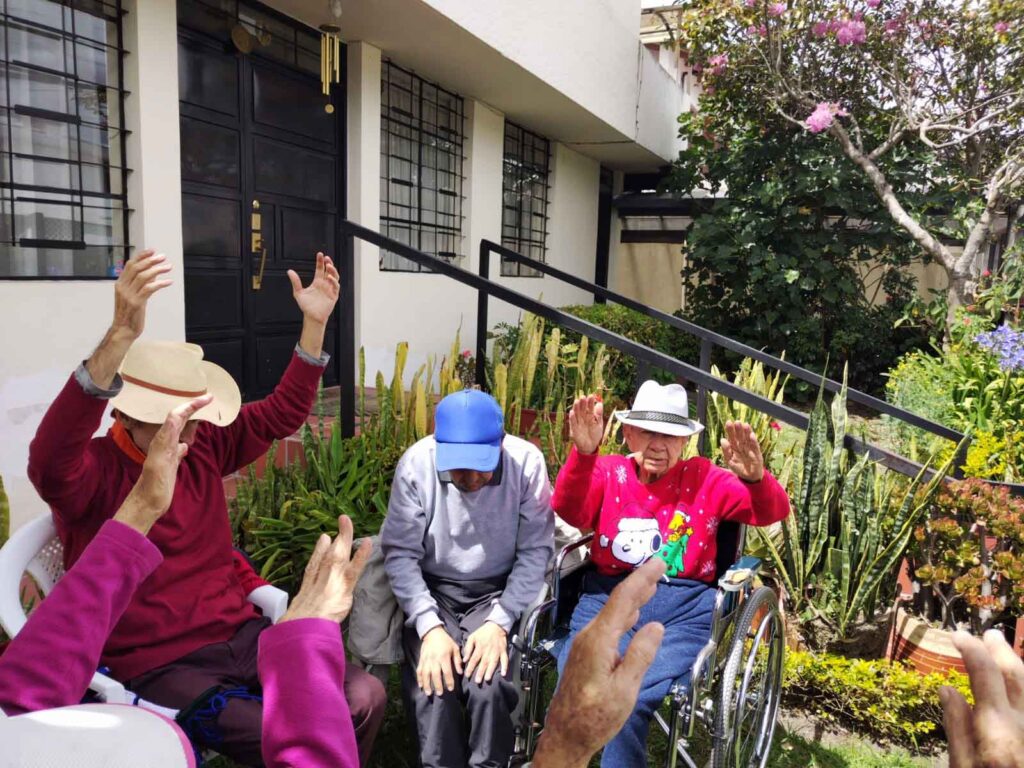 Residentes adultos mayores realizando actividades de estiramiento al aire libre acompañados por personal de enfermería.